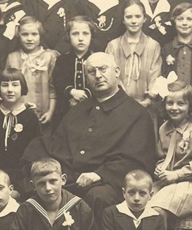 Ein altes Schwarz-Wei├č-Foto eines ernst aussehenden Mannes, der in der Mitte sitzt und von einer Gruppe Kinder umgeben ist, die alle bewundernd zu ihm aufschauen.