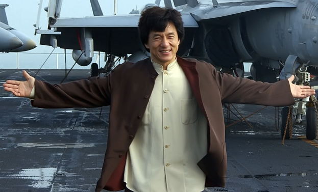 Jackie Chan steht auf dem Deck eines Flugzeugträgers mit ausgebreiteten Armen in einem weißen und braunen Kleid, während Kampfjets und ein klarer blauer Himmel im Hintergrund zu sehen sind.