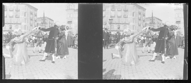 Ein Schwarz-Weiß-Foto einer Gruppe von Menschen, die auf der Straße tanzen, während eines Karnevalsumzugs, mit Kostümen, umgeben von Gebäuden, Straßenlaternen und einer klaren Himmel.