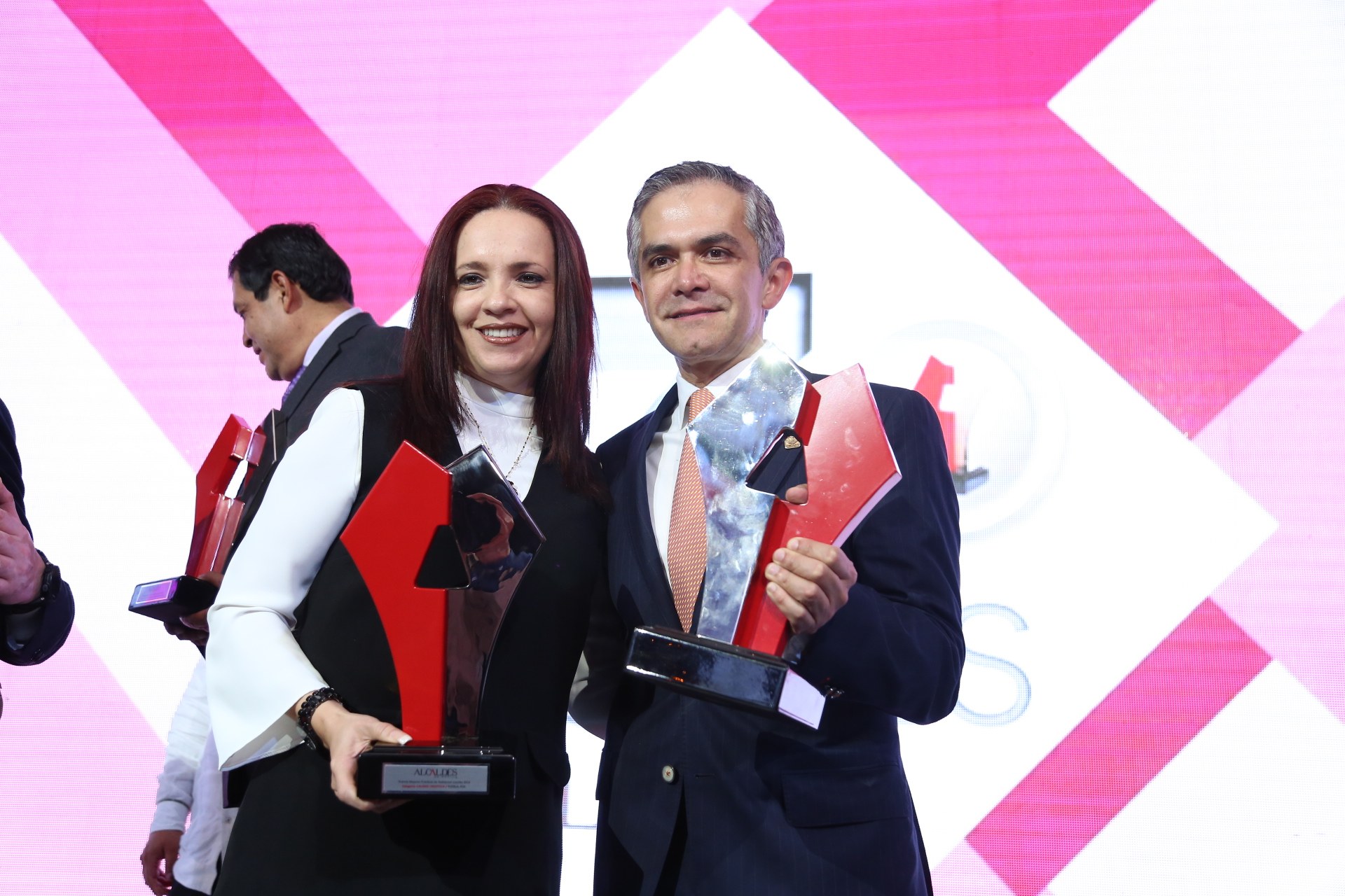 Zwei Personen auf einer Bühne mit Trophäen, die lächeln, mit einem Bildschirm im Hintergrund.