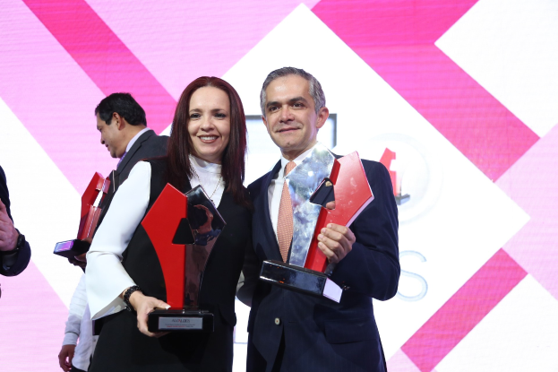 Zwei Personen auf einer Bühne mit Trophäen, die lächeln, mit einem Bildschirm im Hintergrund.