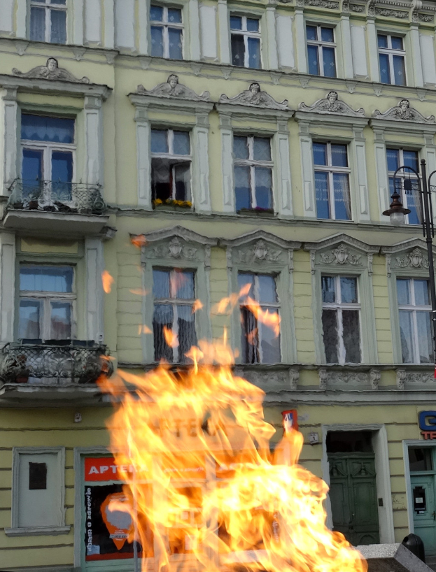 Ein großes Feuer brennt vor einem Gebäude mit Fenstern, Türen, einem Laternenmast und einer Tafel mit einer Inschrift.