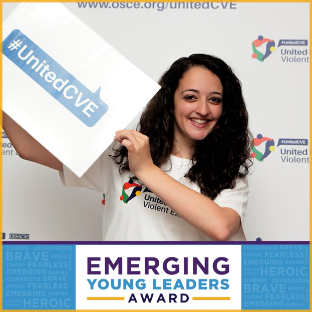 Eine strahlende Frau in einem weißen T-Shirt und Pferdeschwanz hält ein Schild mit der Aufschrift "Emerging Young Leaders Award" hoch und zeigt Freude und Erreichung.