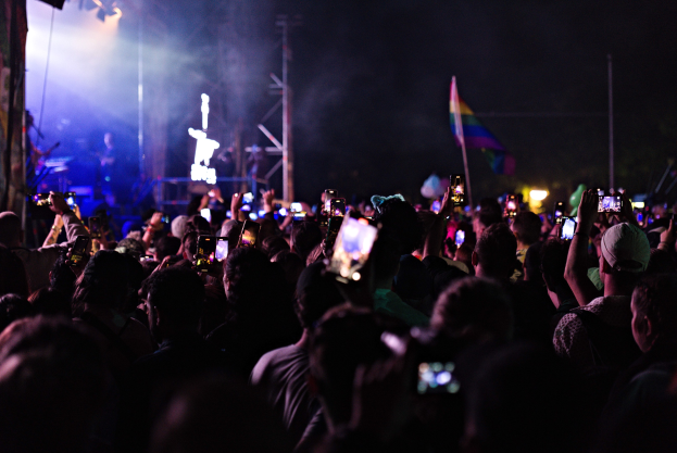 Menge bei einem Konzert mit hochgehaltenen Handys, beleuchtet von Scheinwerfern, umgeben von Pfählen und einer Flagge, während Musiker auf der Bühne spielen.
