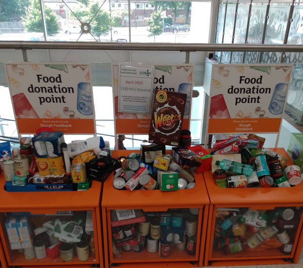 Orangene Schübe mit Lebensmittelspenden wie Flaschen, Dosen und Kisten, mit Informationsschildern und einer Sicht auf Fahrzeuge, Bäume, Gebäude und Himmel durch Glasfenster.