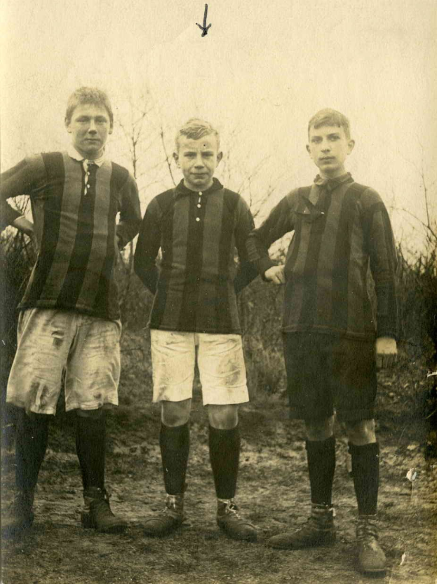 Drei junge Männer in Fußballtrikots stehen Seite an Seite auf einem Rasenfeld mit Bäumen und einem klaren Himmel im Hintergrund.