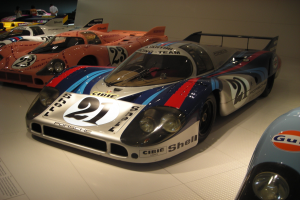 Ein Porsche 917K Le Mans Rennwagen wird in einem Museum ausgestellt, umgeben von anderen Sportwagen, mit Menschen drumherum und einer Wand im Hintergrund und einem Blatt Papier mit Text auf der linken Seite des Bildes.