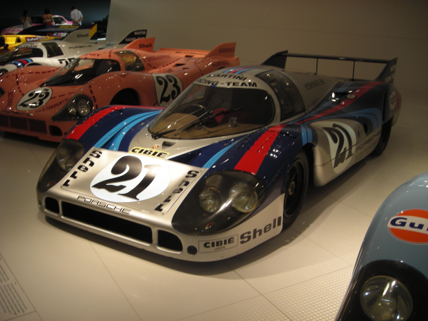 Ein Porsche 917K Le Mans Rennwagen wird in einem Museum ausgestellt, umgeben von anderen Sportwagen, mit Menschen drumherum und einer Wand im Hintergrund und einem Blatt Papier mit Text auf der linken Seite des Bildes.