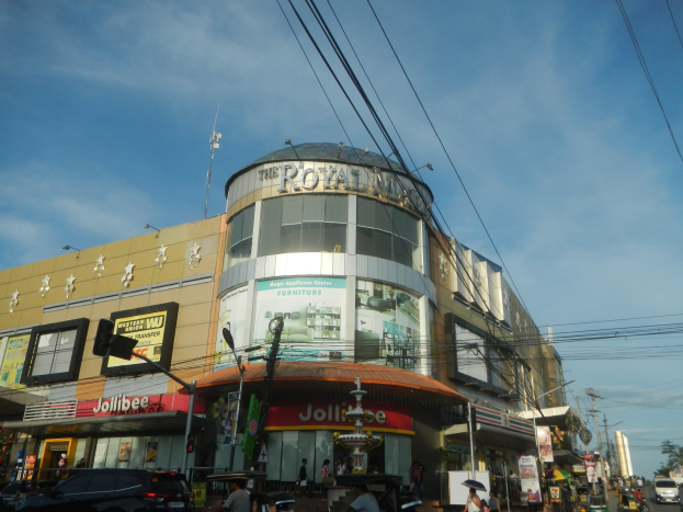 Eine belebte Stadtstraße mit fahrenden Autos, Fußgängern auf dem Gehweg und dem Royal Mall Einkaufszentrum im Hintergrund, umgeben von parkenden Autos, Bäumen, Strommasten und Drähten unter einem klaren Himmel.