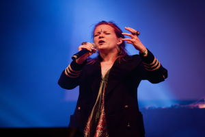 Eine Frau in einem schwarzen Kleid singt in ein Mikrofon auf der Bühne, beleuchtet von blauem Licht, ihre Augen geschlossen und ihr Gesichtsausdruck voller Emotion.