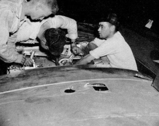 Drei Männer in Mützen reparieren ein schwarz-weißes Auto in einer Garage, umgeben von Werkzeugen und Materialien.