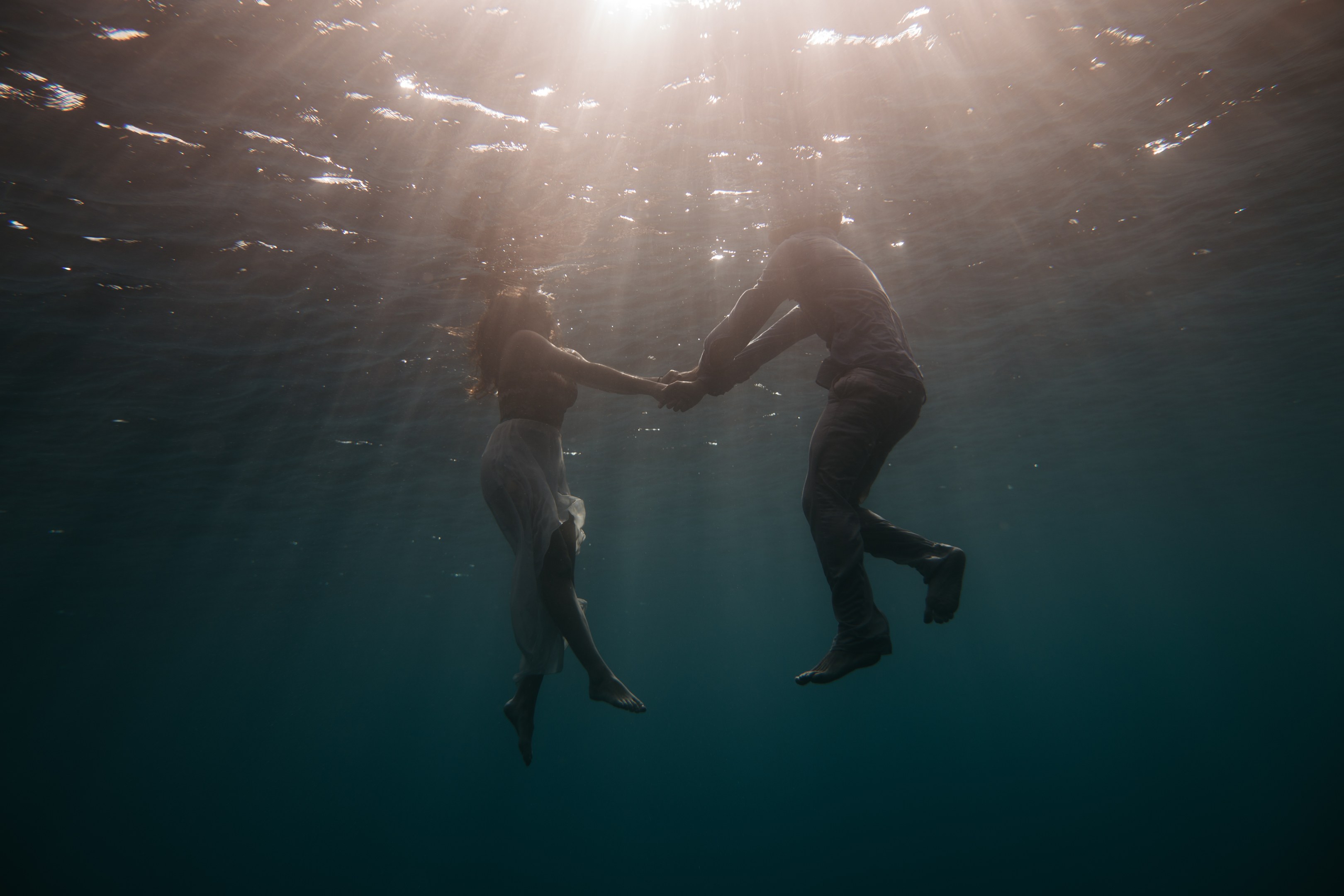 Zwei Personen, die sich unter Wasser in der See an den Händen halten, mit Sonnenstrahlen, die von oben einfallen.