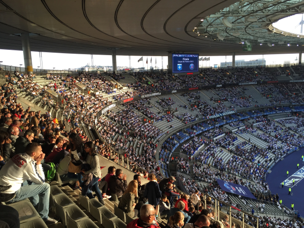 Große Menschenmenge in einem Stadion bei einem Fußballspiel, mit einer Bühne, Fahnen, Pfählen, einem Bildschirm und der Allianz Arena in München, Deutschland im Hintergrund.