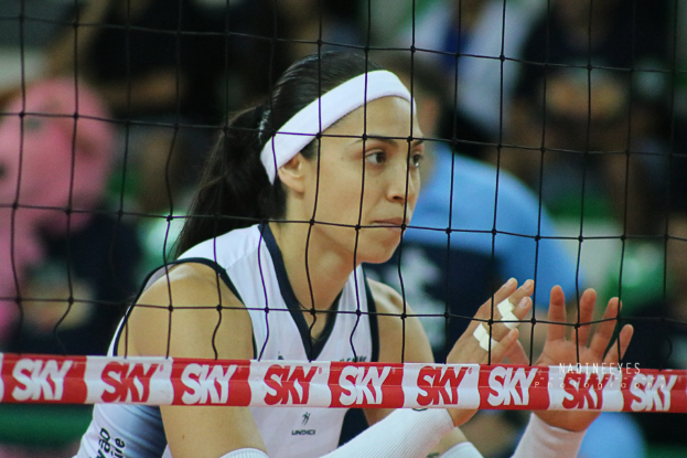 Eine weibliche Volleyballspielerin in Uniform ist im Einsatz auf dem Feld, mit einem Netz im Vordergrund und einem unscharfen Hintergrund, mit einem entschlossenen Gesichtsausdruck.