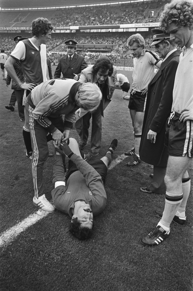 Schwarzes und weißes Bild eines Fußballspielers, der auf dem Boden liegt, während ihn eine Gruppe von Männern und eine Zuschauermenge umgibt, was darauf hindeutet, dass er verletzt sein könnte.