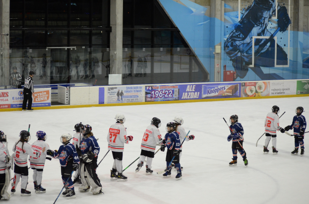 Junge Menschen in Helmen und Eishockeyausrüstung spielen Eisstockschießen auf einer Indoor-Eisbahn mit Bannern, Säulen und Glaswänden im Hintergrund.