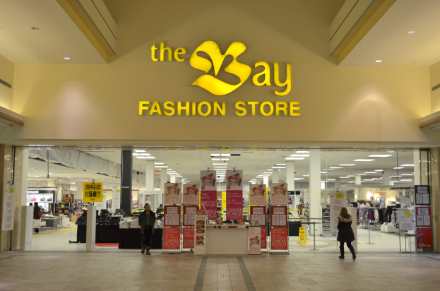 Verkaufsfläche eines Bay Fashion Einzelhandelsgeschäfts in einem Einkaufszentrum mit vorbeilaufenden Kunden, Wandtafeln, Deckenleuchten und Hintergrundauslagen.