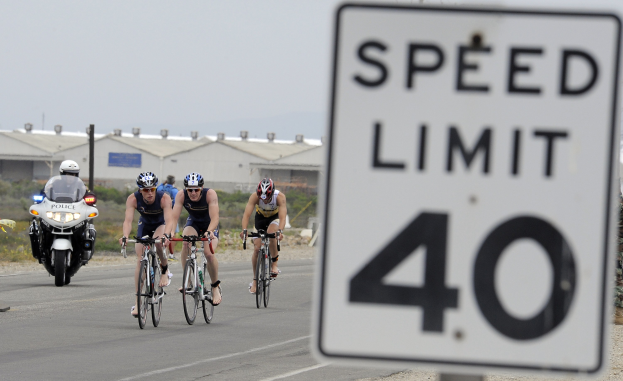 Eine Gruppe von Radfahrern mit Helmen fährt auf einer Straße neben einem Geschwindigkeitsbegrenzungsschild vorbei, mit Gebäuden, Strommasten, Grünflächen und einem klaren blauen Himmel im Hintergrund.