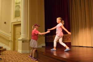 Zwei junge Mädchen tanzen auf einer Bühne vor einem Vorhang, mit einer Person, die links auf einem Stuhl sitzt, einer Säule und einer Wand im Hintergrund, beide Mädchen tragen Schuhe.