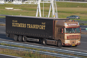 Ein großer Sattelschlepper f√§hrt auf einer Autobahn neben einer Br√ľcke, mit einem Auto daneben, umgeben von Gras, Gel√∂ndern, Pfosten, Brettern und einer Hintergrund von B√§umen, Geb√§uden und einem klaren blauen Himmel.