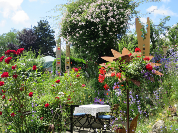 Ein Garten mit bunten Blumen und Pflanzen, mit einem Tisch mit einer Decke und Stühlen, vor Bäumen und einem bewölkten Himmel auf dem Chelsea Flower Show.