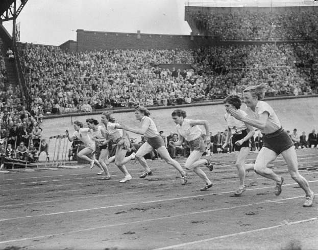 Eine Gruppe von Frauen, die auf einer Laufbahn in einem Stadion laufen, mit Zuschauern auf den Rängen, dargestellt in Schwarz-Weiß.
