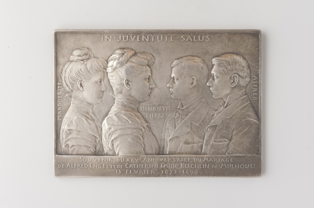 Silberne Plakette mit einem eingravierten Bild von drei Menschen und Text auf einem weißen Hintergrund, wahrscheinlich zur Erinnerung an eine Hochzeit.