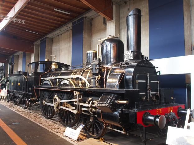 Alte Dampflokomotive in einem Museums-Setting mit umgebenen Eisenbahnschienen, Informationsschildern und anderen Ausstellungsstücken an einer Wand mit Deckenbeleuchtung.