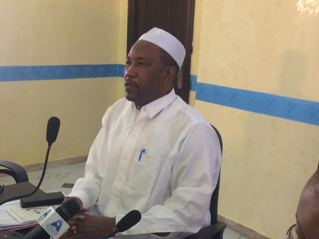 Ein Mann in einem weißen Kleid und Hut sitzt an einem Tisch mit Mikrofonen, spricht, während Papiere und Gegenstände auf dem Tisch sichtbar sind, mit einem anderen Menschen mit Brille rechts von ihm, in einem Raum mit einer Wand und einer Tür im Hintergrund.