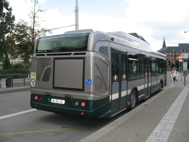 Eine Stadtbus fährt eine Straße mit hohen Gebäuden entlang, auf der rechten Seite ist ein Gehweg mit ein paar Menschen und Polen mit Schildern zu sehen; im Hintergrund sind Bäume, Gebäude und der Himmel zu erkennen.