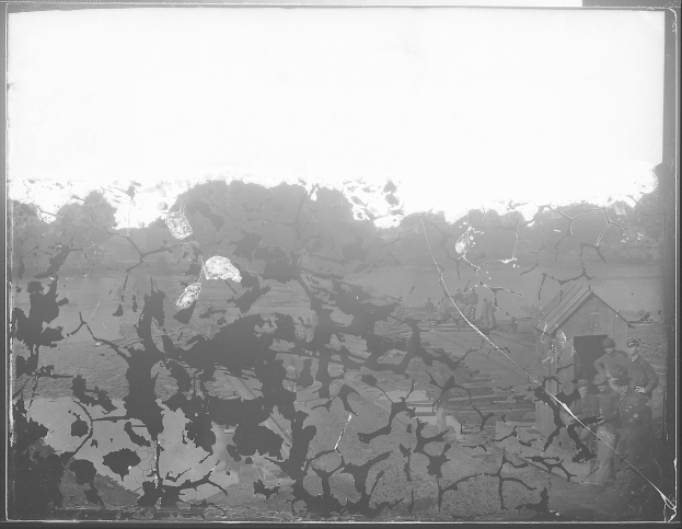 Schwarzes und weißes Foto eines zerbrochenen Fensters mit ein paar Menschen davor, umgeben von Bäumen und einem Haus im Hintergrund.