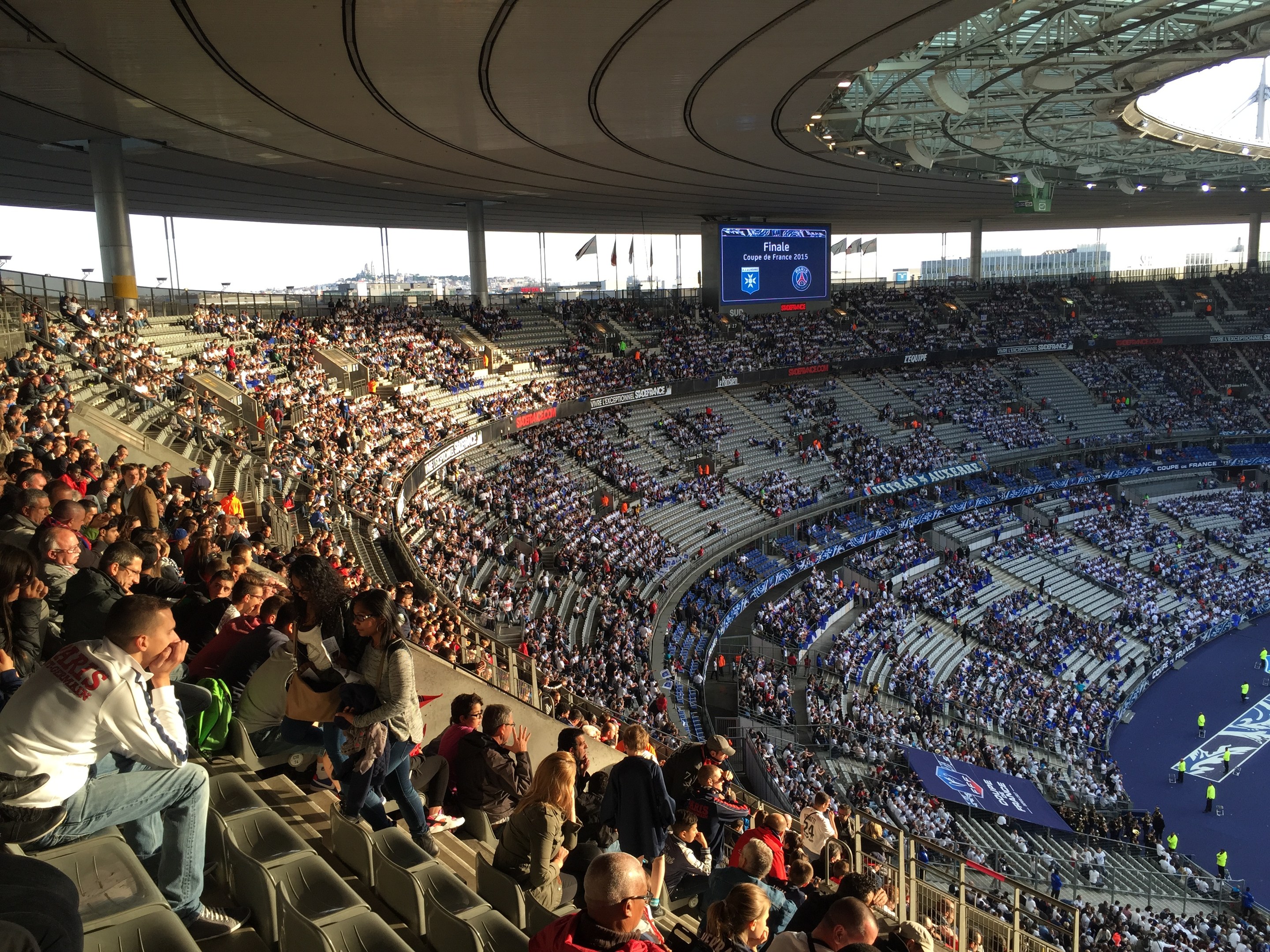Eine große Menschenmenge in einem Stadion schaut ein Fötballspiel zu, mit einer Bühne auf der rechten Seite, Fahnen, Masten, einem Bildschirm und der Allianz Arena in München, Deutschland im Hintergrund.