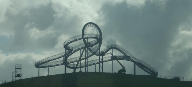 Eine Achterbahn auf dem Höhepunkt eines Hügels mit einem bewölkten Himmel, umgeben von Stützpfählen und -strukturen.