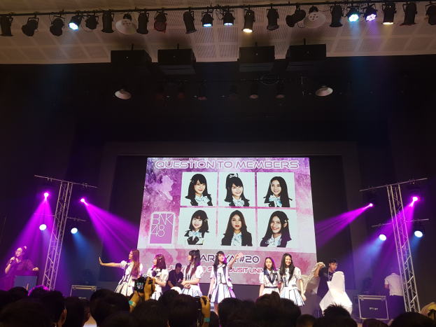 Eine Gruppe von Menschen, die auf einer hell erleuchteten Bühne spielen, mit einer Menge darunter, und einem Menschen mit Mikrofon und einem anderen mit Instrument, während ein großer Bildschirm Bilder von BK48 anzeigt.