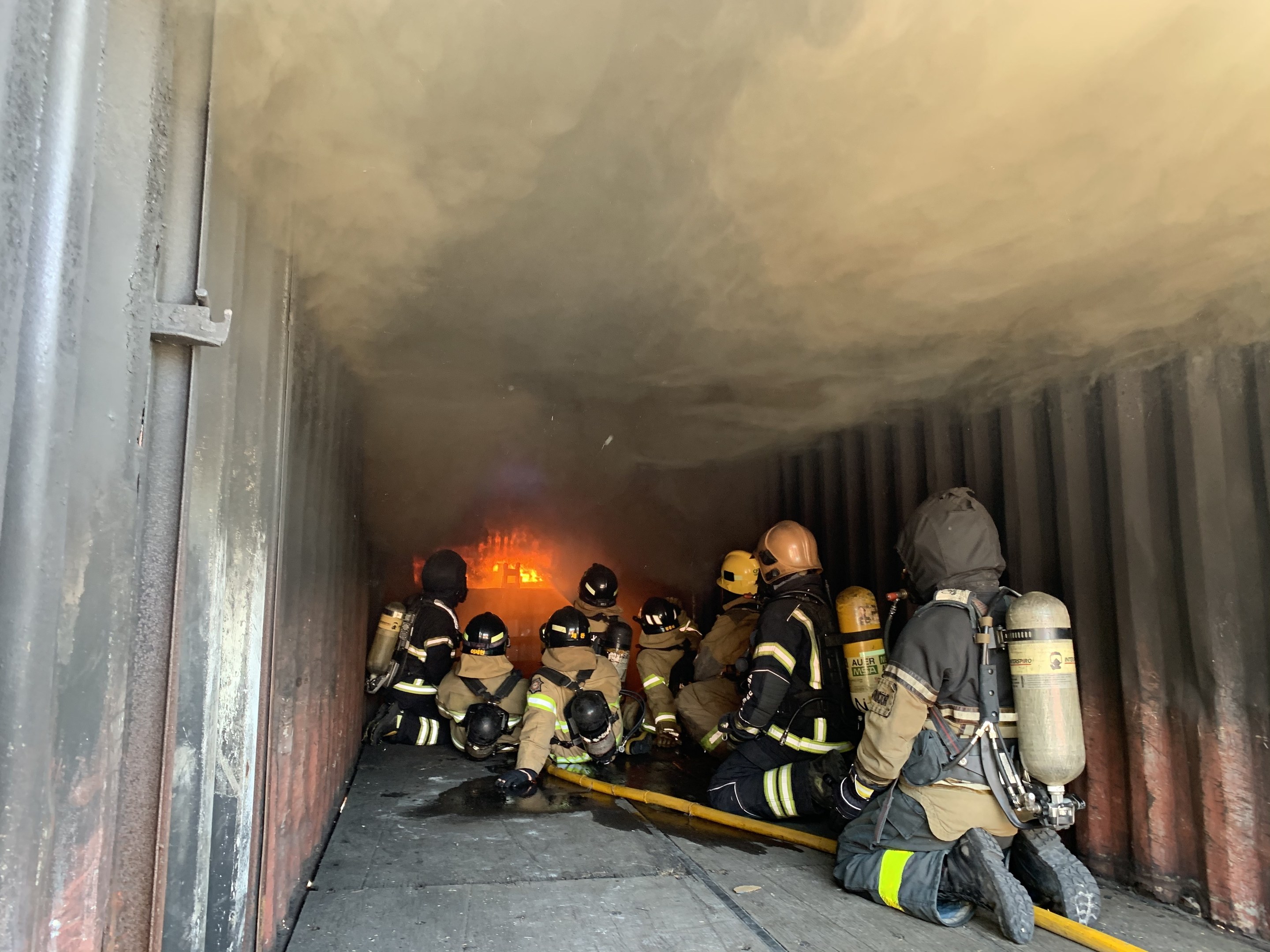 Feuerwehrleute in Schutzanzügen mit Sauerstoffflaschen arbeiten in einem raucherfüllten Tunnel, einer hält einen Schlauch, mit Flammen im Hintergrund und einer Wand auf der linken Seite.