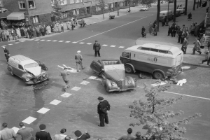 Eine Schwarz-Weiß-Szene eines Autounfalls an einer Straßenkreuzung mit mehreren Fahrzeugen, darunter einem Van, einer Gruppe von Menschen und im Hintergrund Gebäuden, Bäumen, Pfählen und Texttafeln.