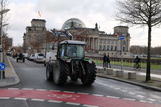 Ein grüner Traktor fährt eine von Bäumen gesäumte Straße vor dem Reichstaggebäude in Berlin, Deutschland, entlang, vorbei an Fußgängern und Radfahrern auf dem Gehweg, Laternen, Schildern und Fahnen auf dem Gebäude bei einem bewölkten Himmel.