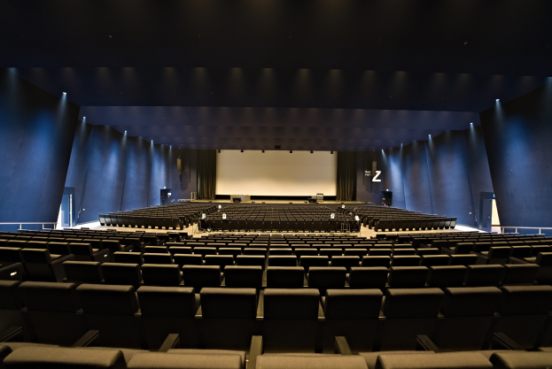 Großer Zuschauerraum mit Sitzreihen, Projektionsschirm und Bühnenobjekte, beleuchtet von Wandlampen.