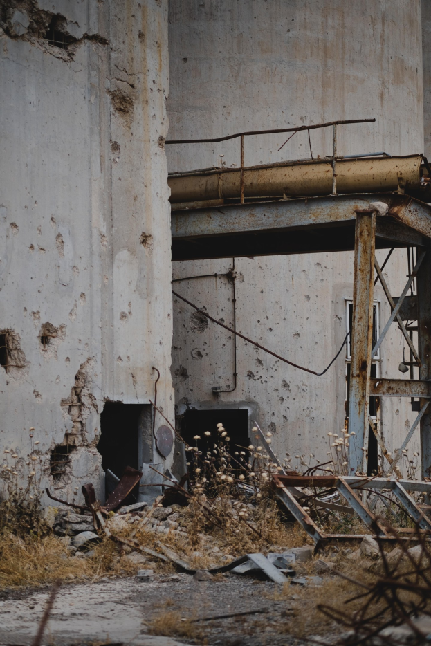 Altes Gebäude mit einem zentralen Metallgerüst, umgeben von Pflanzen, Eisenstangen und Trümmern, mit bröckelnden Wänden und zerbrochenen Fenstern.