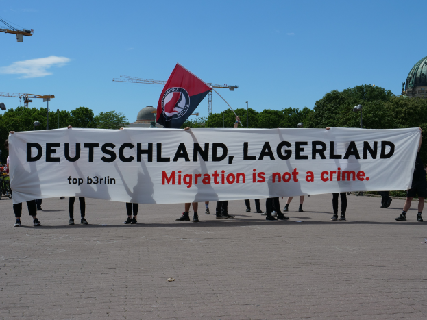 Eine Gruppe von Menschen hält eine Fahne mit der Aufschrift "Deutschland, Lagerland Migration ist kein Verbrechen" und eine Flagge, die auf dem Boden steht mit Straßenlaternen, Bäumen, Kränen, einem Gebäude und einem bewölkten Himmel im Hintergrund.