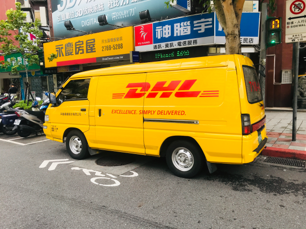 Ein gelber DHL-Lieferwagen steht am Straßenrand neben Gebäuden, Bäumen, Schildern, Pfählen, Verkehrszeichen und einer Person auf dem Gehweg.