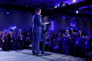 Ein Mann steht an einem Podium mit einem Mikrofon und spricht zu einer Menge, während einige klatschen und Schilder halten, unter Deckenlampen bei einer Wahlveranstaltung.