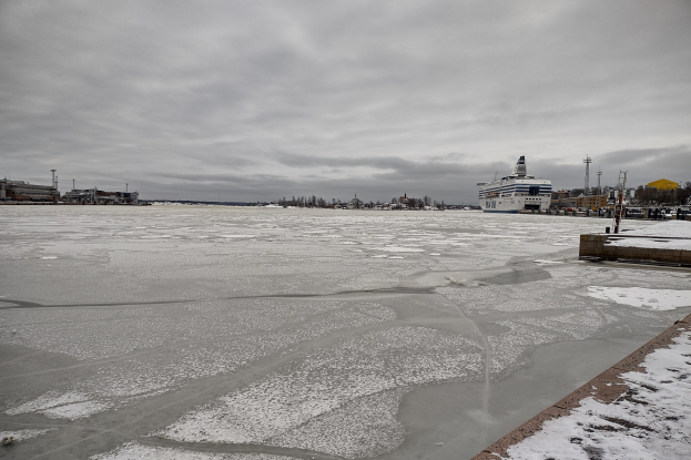 Großes Kreuzfahrtschiff liegt nahe einem schneebedeckten Dock mit Gebäuden, Bäumen und Masten im Hintergrund unter einem bewölkten Himmel.