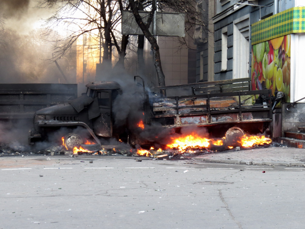 Ein gro├če Lkw brennt an der Stra├če mit dickem Rauch, der in die H├Âhe steigt, B├Ąume und Geb├Ąude sind im Hintergrund unter einem klaren Himmel zu sehen.