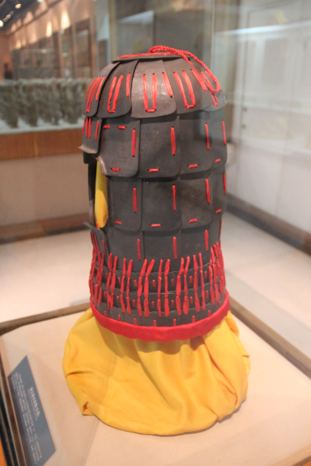 Helm in einer Glasvitrine mit einem gelben Tuch darauf, begleitet von einer Texttafel auf der linken Seite und Deckenleuchten im Hintergrund.