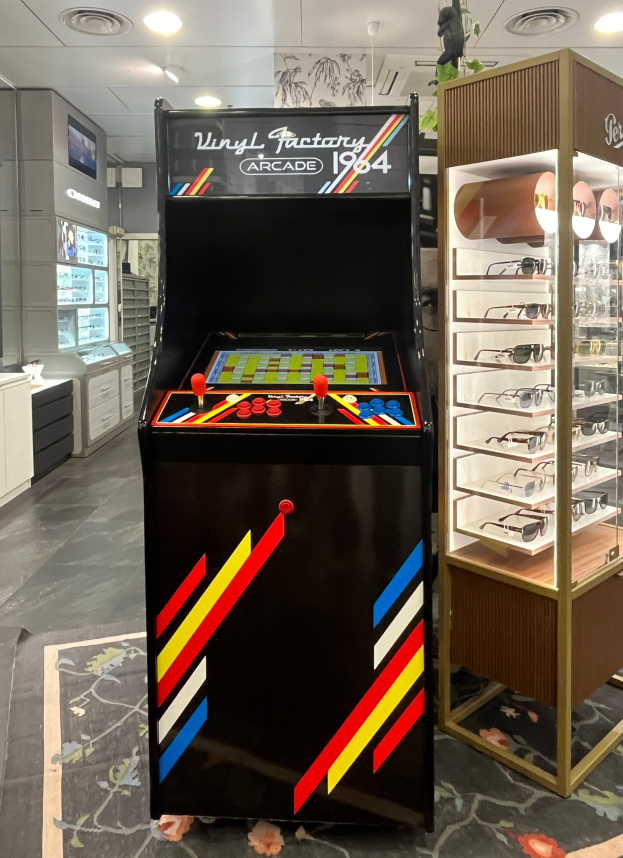 Ein vintage Arcade-Automat steht in der Mitte eines retro-stilvollen Ladens mit einer Brillenablage auf der rechten Seite, Schränken, einem Fernseher und Beleuchtung im Hintergrund.