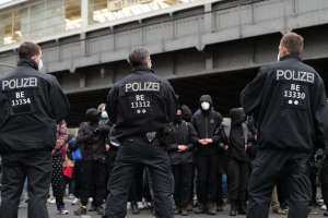 Eine Gruppe von Polizisten in Uniform steht vor einer Menge von Menschen in schwarzen Uniformen und Masken, mit einer Brücke und einem Gebäude im Hintergrund, was auf eine städtische Umgebung hindeutet.