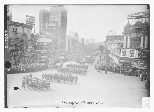 Ein Schwarz-Weiß-Foto eines Umzugs in einer Stadt, mit Menschen, die die Straße entlangmarschieren, einige mit Gewehren, und Zuschauern an den Seiten, mit Gebäuden und Pfählen im Hintergrund, und Text unten.