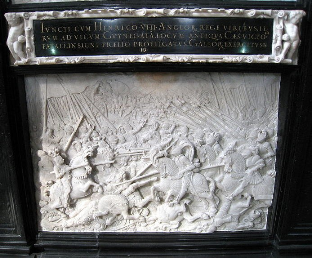 Eine weiße Marmorplatte mit schwarzem Rahmen an einem Gebäude zeigt eine Kampfszene mit Schwert- und Schildträgern, umgeben von Zuschauern.