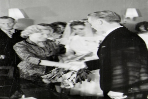 Ein Schwarz-Weiß-Foto einer Braut in einem weißen Kleid und Schleier, die einen Strauß hält, und eines Bräutigams in einem Anzug und Krawatte, die Ringe tauschen, mit Lampen im Hintergrund.